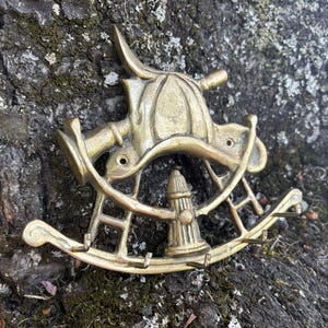 Könnte beinhalten: Ein messingfarbener Schlüsselhalter in Form eines Schaukelpferds mit einem Feuerwehrhelm- und Hydranten-Design. Der Schlüsselhalter hat drei Haken und ist auf einer strukturierten, dunklen Oberfläche montiert.