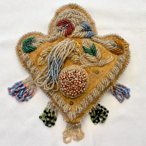 Op de afbeelding: Een hartvormig tasje van beige leer, versierd met ingewikkeld kralenwerk in zilver, rood, blauw, groen en geel. Het oppervlak is voorzien van bloemmotieven, een centrale rozet van kralen en bungelende kralenkwastjes in diverse kleuren.