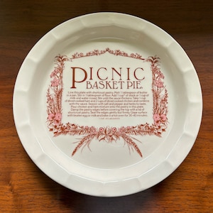Puede incluir: Plato para tartas de cerámica blanca con un borde floral marrón y rosa y el texto "PICNIC BASKET PIE." El texto incluye una receta para una tarta de cesta de picnic.