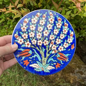 May include: A round, hand-painted ceramic plate with a vibrant blue background. The plate features a stylized tree design with white flowers, turquoise leaves, and red and orange tulip-like accents. The plate is held by a hand.