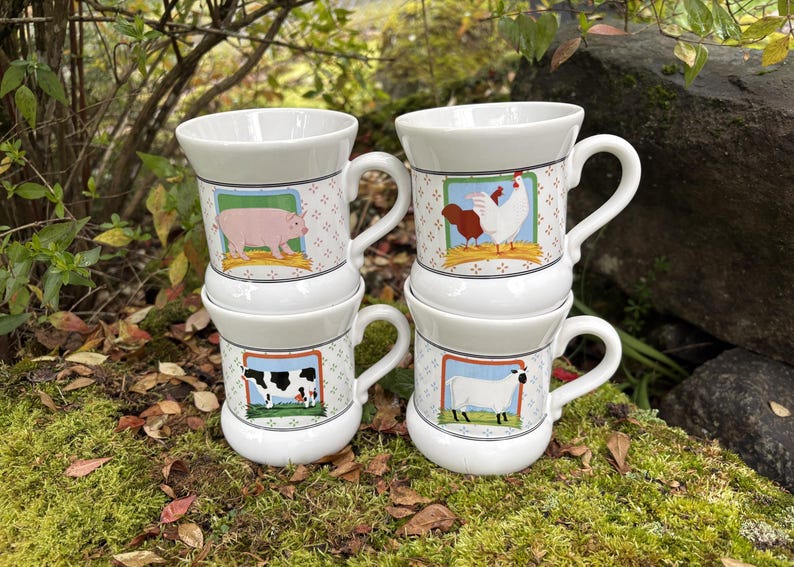 May include: Four white ceramic mugs stacked, each with a different farm animal illustration: a pig, a rooster and hen, a cow, and a sheep. The mugs have a patterned design and are set on a bed of moss.