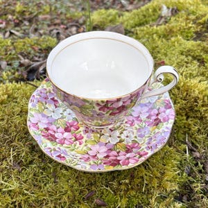 May include: A floral teacup and saucer set. The teacup and saucer feature a pattern of purple, pink, and white flowers with green leaves. The teacup has a gold rim and handle. The set is placed on a bed of green moss.