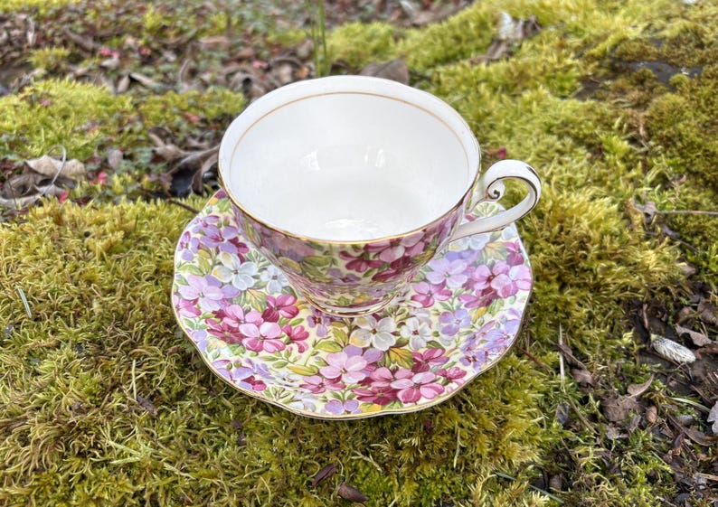 May include: A floral teacup and saucer set. The teacup and saucer feature a pattern of purple, pink, and white flowers with green leaves. The teacup has a gold rim and handle. The set is placed on a bed of green moss.