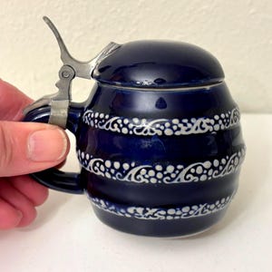 以下が含まれることがあります： ヒンジ付きの金属製の蓋とハンドルが付いた、小さな濃い青色のセラミックビールジョッキ。ジョッキは、白く装飾的なスクロールのような模様で装飾されています。ジョッキは手で持たれています。