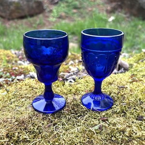 Puede incluir: Dos copas de vino vintage azul cobalto. Una tiene un cuenco acampanado y una base de pedestal, la otra un patrón en relieve. Las copas se exhiben sobre musgo verde. Las copas son de un llamativo tono azul.