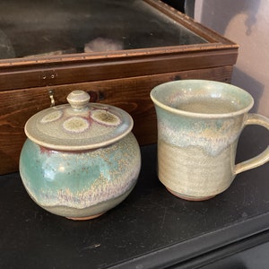 Peut inclure: Un pot en céramique vert et brun avec un couvercle et une tasse en céramique vert et brun. Le pot a un glaçage moucheté et la tasse a un glaçage moucheté.
