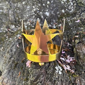 Puede incluir: Una corona de metal dorado con recortes y puntas de estrella. La corona est&aacute; sobre una superficie texturizada de color marr&oacute;n oscuro con peque&ntilde;as hojas y musgo.