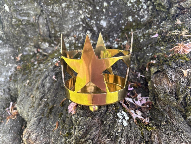 Puede incluir: Una corona de metal dorado con recortes y puntas de estrella. La corona est&aacute; sobre una superficie texturizada de color marr&oacute;n oscuro con peque&ntilde;as hojas y musgo.