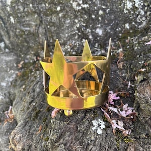 Puede incluir: Un portavelas de metal dorado con forma de corona, con recortes de estrellas. El portavelas cil&iacute;ndrico tiene una base redondeada y est&aacute; sobre un tronco de &aacute;rbol.
