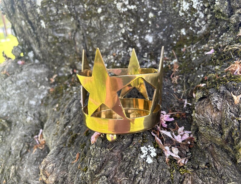 Puede incluir: Un portavelas de metal dorado con forma de corona, con recortes de estrellas. El portavelas cil&iacute;ndrico tiene una base redondeada y est&aacute; sobre un tronco de &aacute;rbol.