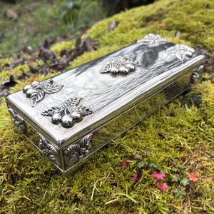 May include: A rectangular silver box with ornate leaf and berry designs on the lid and corners. The box has a wood-grain texture on the top and is set against a backdrop of green moss and foliage.