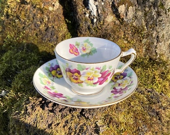 May include: A white teacup and saucer with a floral pattern of purple, yellow, and white pansies. The cup and saucer have a gold trim.