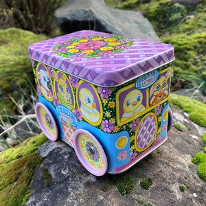 May include: A vintage tin box shaped like a bus with a pastel pink, blue, and green floral design. The bus has the words "Easter Express" on the side and features cartoon animals like a bunny, chick, and lamb.