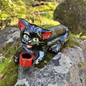 May include: A black ceramic cat figurine with green eyes and red ear accents. The cat is holding a red bowl. The cat's back has a striped design. The figurine is resting on a moss-covered rock.