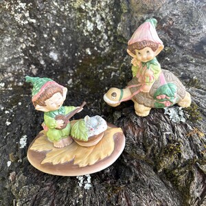 Könnte beinhalten: Zwei Keramikfiguren, die kleine elfenartige Wesen darstellen. Ein Elf trägt eine grüne Mütze und spielt ein Saiteninstrument neben einem kleinen Korb. Der andere Elf trägt eine rosa blütenförmige Mütze und sitzt auf einer braunen Schildkröte.