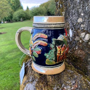 Può includere: Una boccale di birra in ceramica blu e marrone con manico e un design a rilievo di spighe di grano, fiori e cavalli. La boccale è appoggiata su un tronco d'albero.