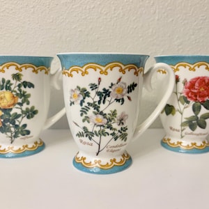 Op de afbeelding: Drie witte keramische mokken met bloemmotieven. Elke mok heeft een andere rozenillustratie, met een turquoise rand en basis en gouden rand. De mokken hebben gebogen handvatten.