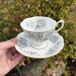 May include: White teacup and saucer set with a gray leaf pattern and gold trim. The teacup has a curved handle and sits on a matching saucer. The set is held in front of a blurred green and red background.