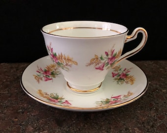 May include: A white teacup and saucer with a floral pattern. The cup and saucer have a gold trim around the edges. The flowers are pink and yellow with green leaves.
