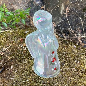May include: A white iridescent glass angel figurine with painted red berries and white flowers. The angel is praying with its hands clasped together.