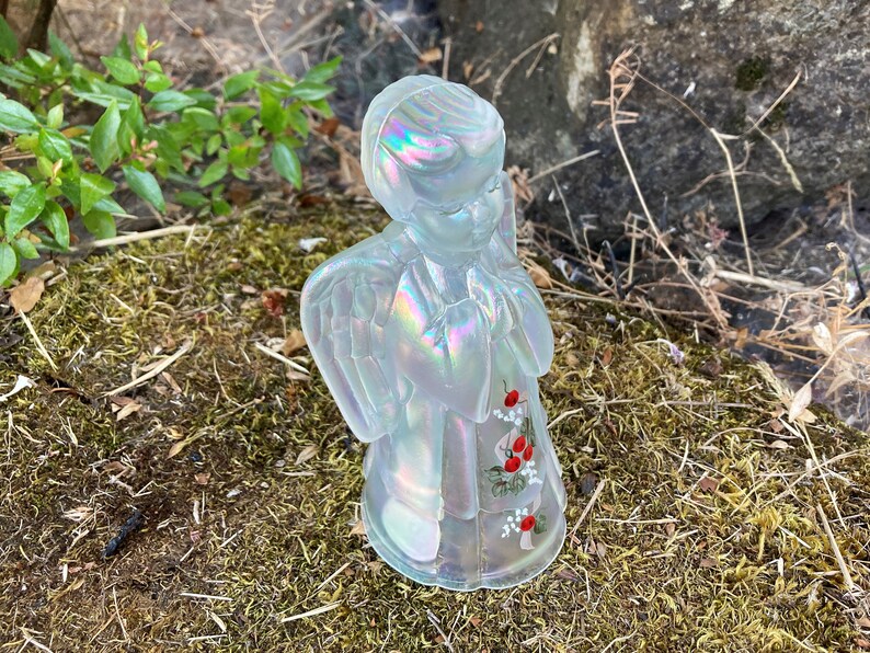 May include: A white iridescent glass angel figurine with painted red berries and white flowers. The angel is praying with its hands clasped together.