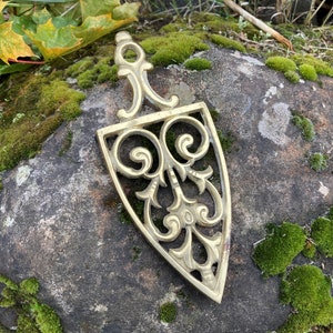 May include: A gold-colored metal trivet with a decorative, ornate design. The trivet is shaped like a pointed oval and has a loop at the top for hanging.