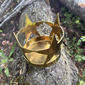 Puede incluir: Un portavelas de metal dorado con dise&ntilde;o de estrella. El portavelas tiene una base circular y est&aacute; adornado con recortes en forma de estrella y detalles de estrella. El objeto est&aacute; sobre un tronco de &aacute;rbol.