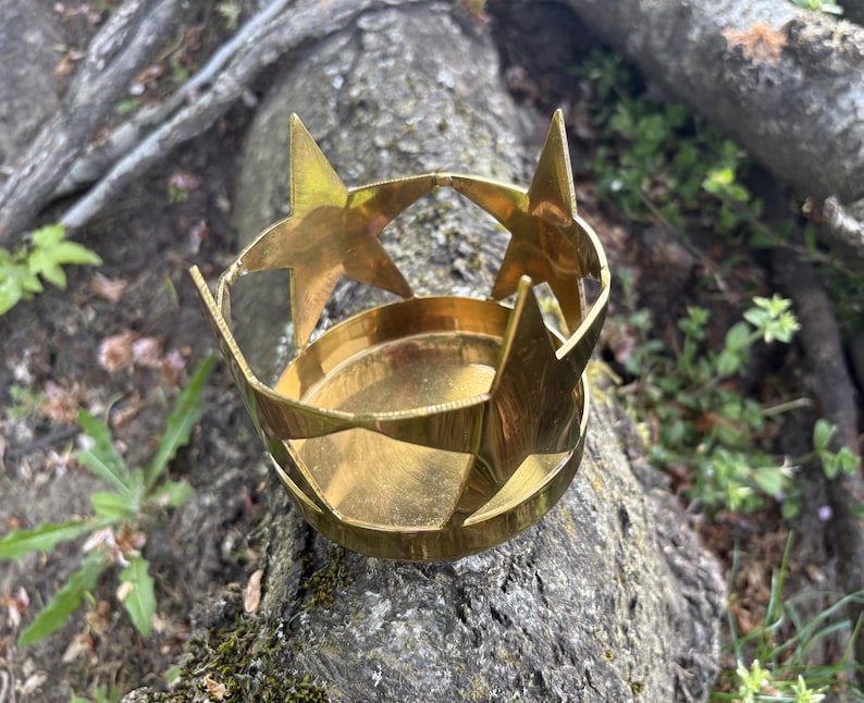 Puede incluir: Un portavelas de metal dorado con dise&ntilde;o de estrella. El portavelas tiene una base circular y est&aacute; adornado con recortes en forma de estrella y detalles de estrella. El objeto est&aacute; sobre un tronco de &aacute;rbol.