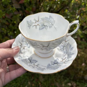 May include: White teacup and saucer set with a delicate gray leaf pattern and gold trim. The teacup has a curved handle and sits on a matching saucer. The set is made of porcelain and is ideal for serving tea.