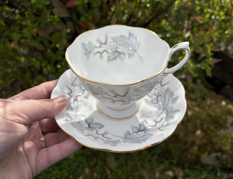 May include: White teacup and saucer set with a delicate gray leaf pattern and gold trim. The teacup has a curved handle and sits on a matching saucer. The set is made of porcelain and is ideal for serving tea.