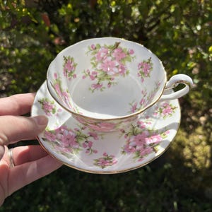 K&ouml;nnte beinhalten: Wei&szlig;es Tee-Set mit rosa Blumenmuster und goldenem Rand. Die Tasse wird in einer Hand gehalten, wodurch das florale Design hervorgehoben wird. Ideal f&uuml;r Tee, dieses Set verleiht jedem Anlass einen Hauch von Eleganz.