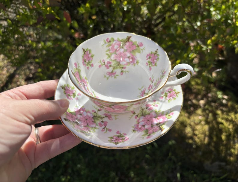 K&ouml;nnte beinhalten: Wei&szlig;es Tee-Set mit rosa Blumenmuster und goldenem Rand. Die Tasse wird in einer Hand gehalten, wodurch das florale Design hervorgehoben wird. Ideal f&uuml;r Tee, dieses Set verleiht jedem Anlass einen Hauch von Eleganz.