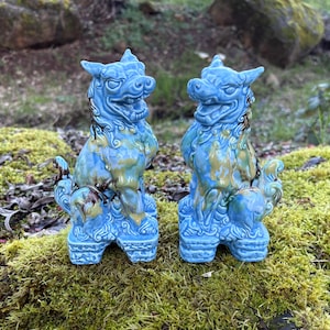 May include: Two blue ceramic Foo Dog statues with detailed features and a glossy finish. The statues have a mix of blue, yellow, and brown colors. They are standing on a bed of green moss.