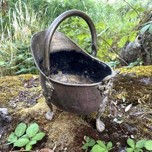 May include: A small, antique-style metal coal scuttle with a dark, weathered patina. It features a high, arched handle, a deep basin with a dark interior, and three ornate, claw-shaped feet attached to the sides with decorative metal mounts.