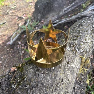 Puede incluir: Una corona dorada en forma de estrella de cinco puntas, apoyada sobre un tronco de &aacute;rbol texturizado y cubierto de musgo. La corona est&aacute; hecha de metal plegado y tiene una base circular. El fondo muestra un entorno natural borroso.