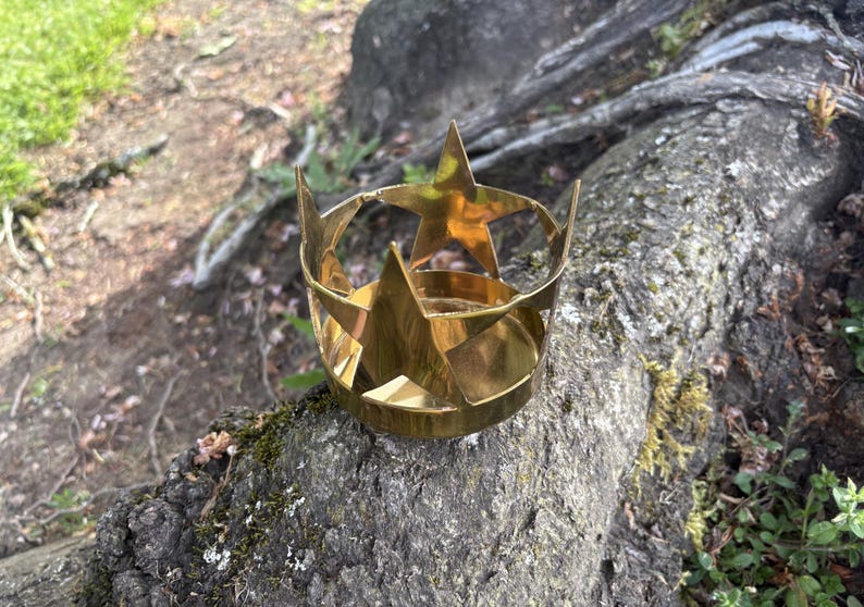 Puede incluir: Una corona dorada en forma de estrella de cinco puntas, apoyada sobre un tronco de &aacute;rbol texturizado y cubierto de musgo. La corona est&aacute; hecha de metal plegado y tiene una base circular. El fondo muestra un entorno natural borroso.