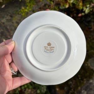 May include: A white Royal Albert saucer with a scalloped edge. The underside features a gold emblem with text that reads "Royal Albert, Bone China, England, Silver Maple."