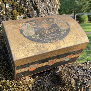 Könnte beinhalten: Eine antike Whitman's Chocolates Blechdose mit gold-schwarzem Design. Der Deckel zeigt drei Wappen und den Text "Whitman's Prestige Chocolates".