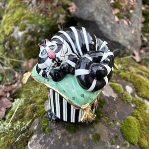 Könnte beinhalten: Eine skurrile Keramik-Katzenfigur mit schwarz-weißen Streifen, die auf einem grünen Kissen mit goldenem Rand ruht. Die Katze hat rosa Ohren und grüne Augen. Der Sockel ist schwarz-weiß gestreift.