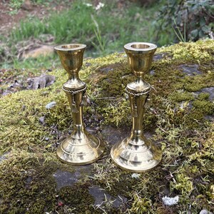 May include: Two antique brass candle holders with a decorative rope and tassel design. The candle holders are displayed on a mossy surface, with a blurred background of greenery.