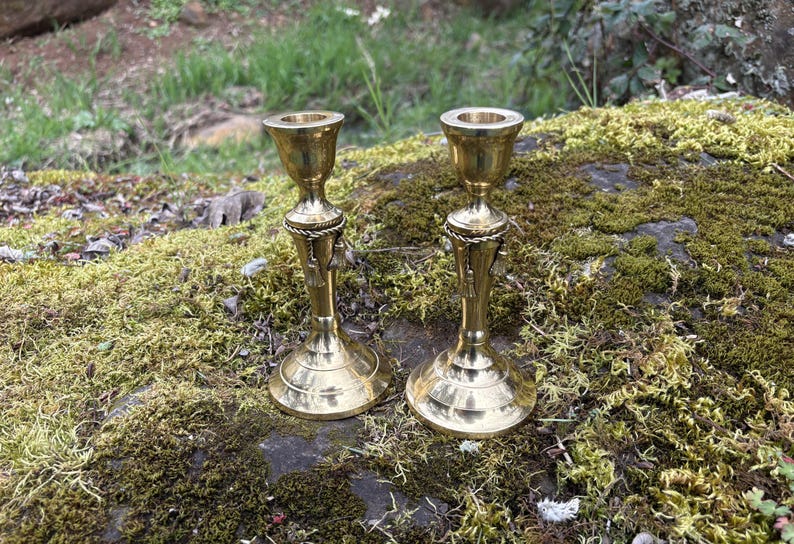 May include: Two antique brass candle holders with a decorative rope and tassel design. The candle holders are displayed on a mossy surface, with a blurred background of greenery.