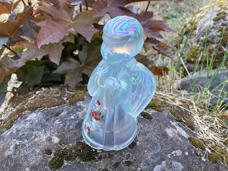 May include: A white iridescent glass angel figurine with painted red berries and white flowers. The angel is standing with its hands clasped in prayer and has wings.