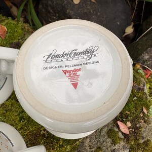 May include: White ceramic mug with the text "Vandor Country Collection" and "Designer: Pelzman Designs". The mug is marked "Made in Japan" and "&copy;1981". The mug is sitting on a bed of green moss.