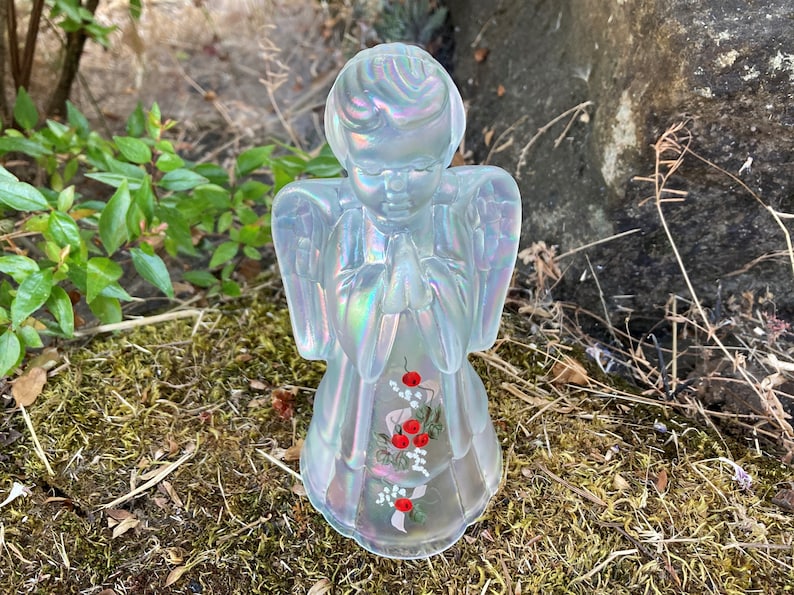 May include: A clear glass angel figurine with iridescent wings and painted red berries and white flowers. The angel is praying with its hands clasped together.