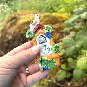 May include: A small ceramic cuckoo clock ornament featuring a white bird perched on a brown roof, green leaves, and blue grapes. The clock face is white with black numbers and hands, set against a light brown wooden-textured frame with painted details.