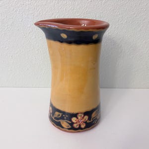 May include: A ceramic pitcher with a flared rim and a curved body. The pitcher has a tan body with a dark blue band at the top and bottom, decorated with floral and leaf motifs. The rim is a reddish-brown color.