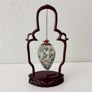 May include: A decorative, dark red wooden stand with an arched top, holding a glass egg-shaped ornament. The ornament features a painted landscape with trees and is suspended by a chain. The base of the stand has carved details.