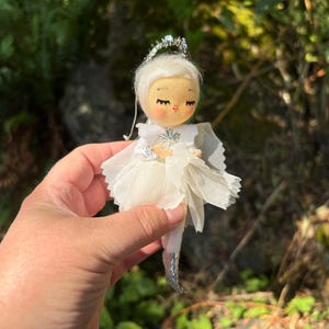 May include: A small doll ornament featuring a white felt face with closed eyes, pink cheeks, and white hair. The figure wears a layered white sheer fabric dress with a silver star accent at the chest, a silver halo, and silver textured pointed boots.