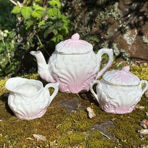 以下が含まれることがあります： 白とピンクの磁器ティーセット。ティーポット、クリーマー、シュガーボウルが含まれています。セットは花の模様と金のトリムが特徴です。ティーポットには蓋と曲がった注ぎ口が付いています。