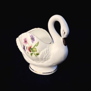 May include: A white porcelain swan-shaped dish featuring a delicate floral pattern of purple and red violets on its side. The swan has a curved neck, a gold-painted beak, and a gold-trimmed rim along the base and the top opening of the vessel.